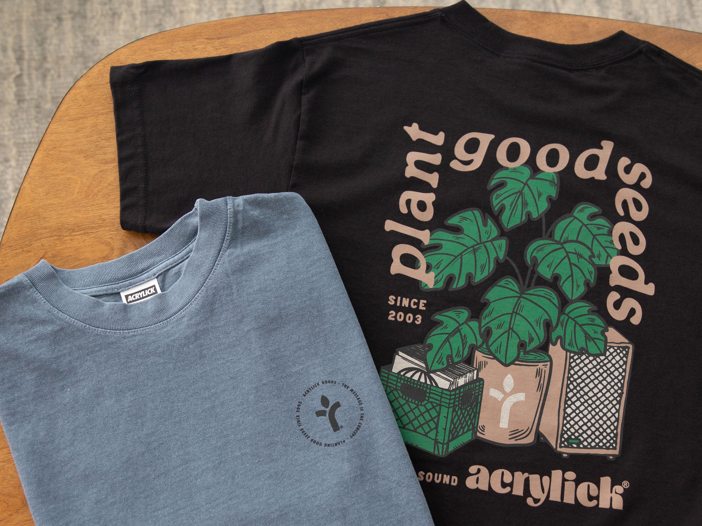 Two t-shirts, one black with a plant-themed design and one blue with a logo, on a wooden surface.