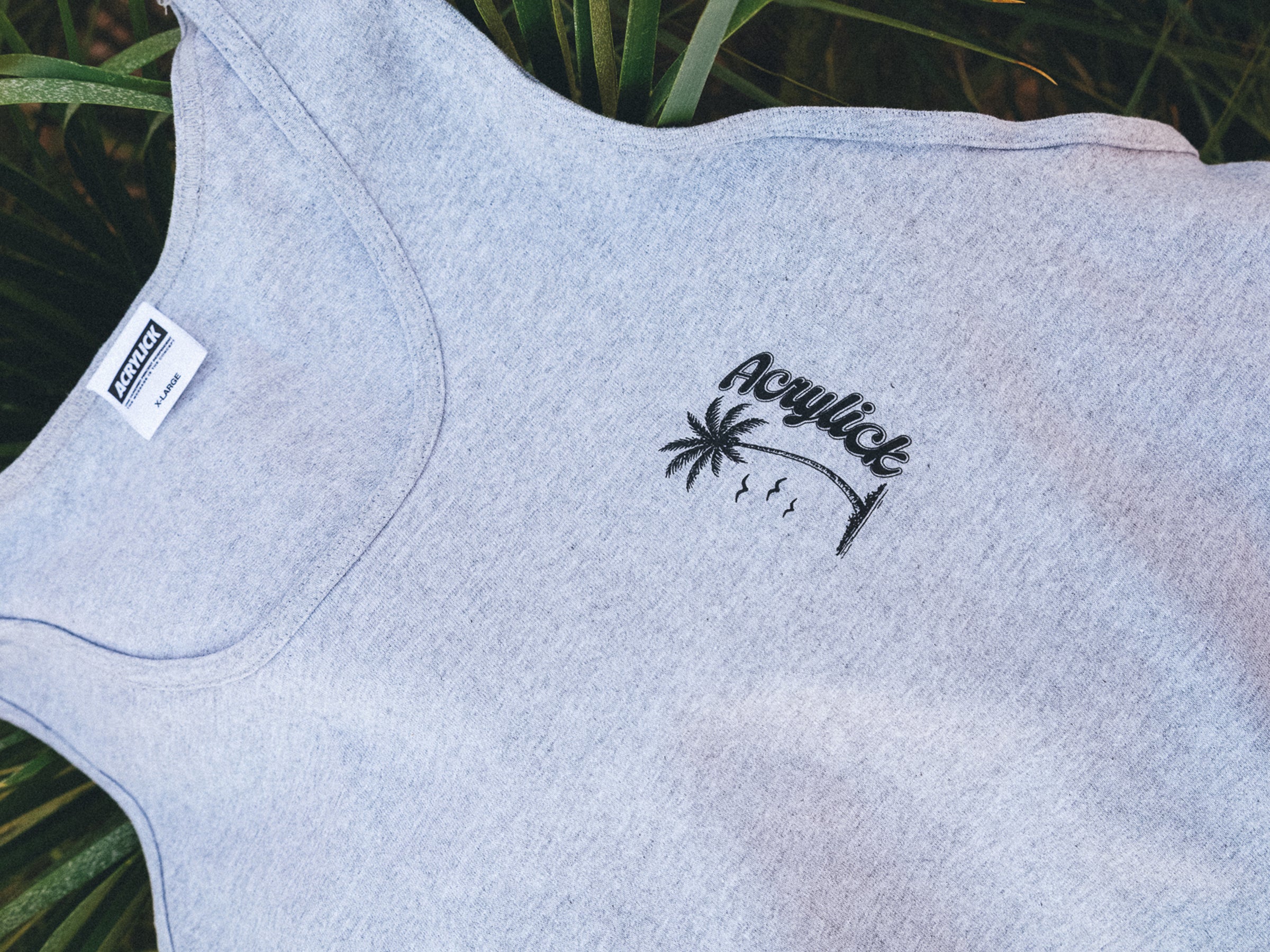Gray tank top with a palm tree design and 'Acrylick' branding on a grassy background